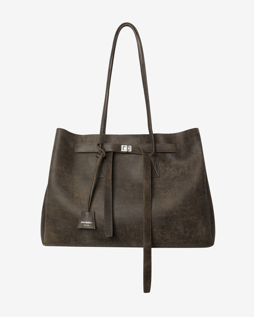 A dark brown distressed leather tote bag with long shoulder straps, a front tie detail, and a small metal logo plaque, shown against a plain light gray background.