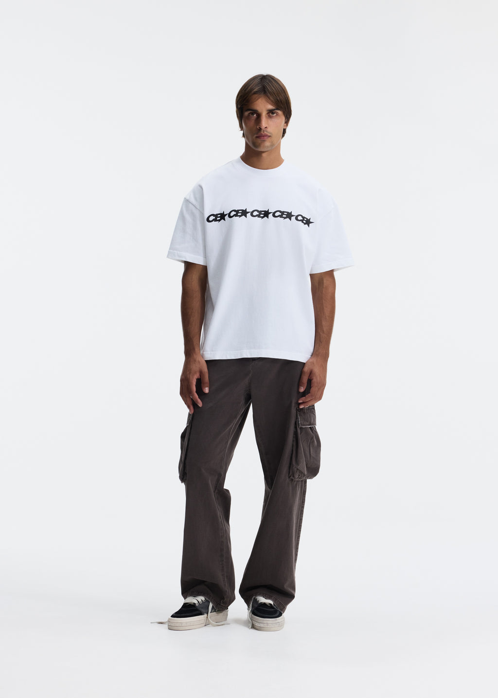 A male model stands against a plain white studio background wearing a white short-sleeve T-shirt with black text across the chest, loose dark cargo pants, and black sneakers.