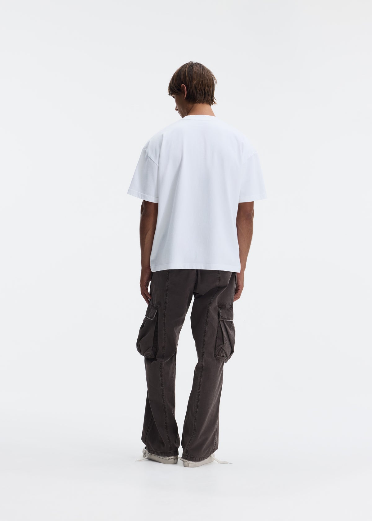 A model shown from the back wearing a white oversized short-sleeve t-shirt and dark brown wide-leg cargo pants, standing against a seamless white studio background.