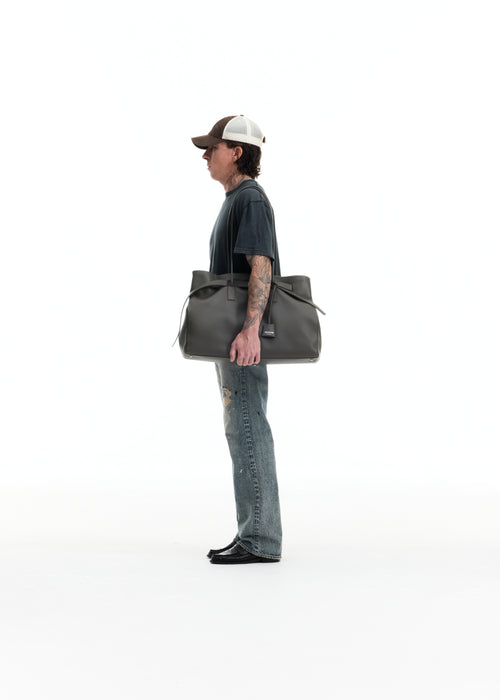 A man in a side profile wearing a dark t-shirt, blue jeans, and a trucker hat, carrying a large dark gray leather tote bag over his shoulder against a solid white background.
