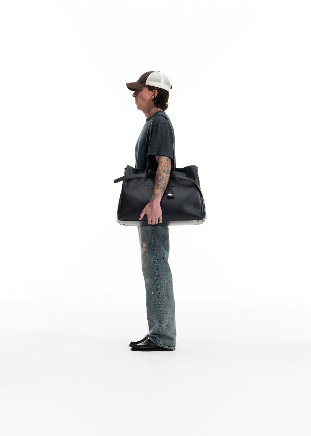 A man in a side profile wearing a baseball cap, dark t-shirt, and jeans, carrying a large black leather tote bag against a solid white background.