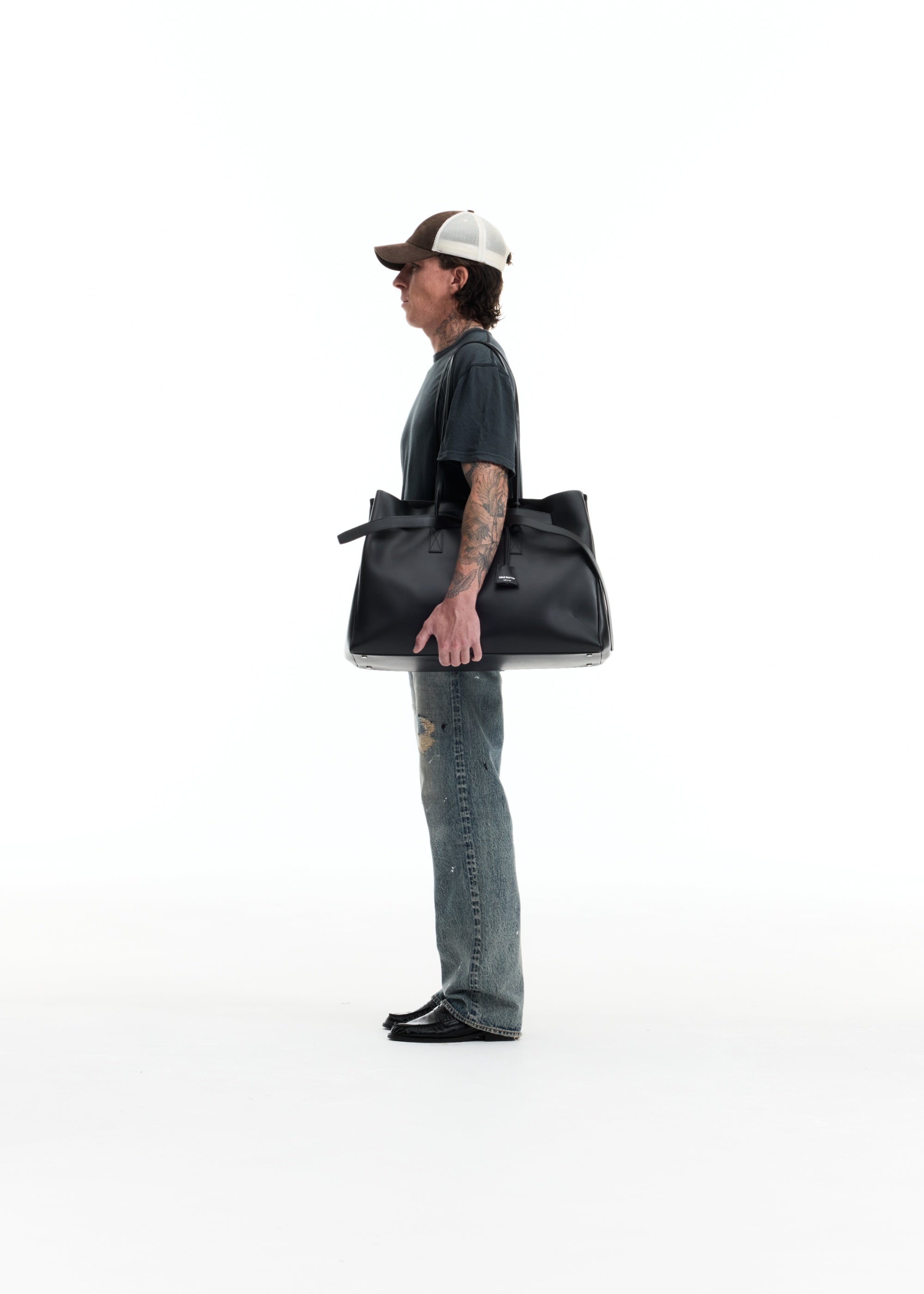 A man in a side profile wearing a baseball cap, dark t-shirt, and jeans, carrying a large black leather tote bag against a solid white background.
