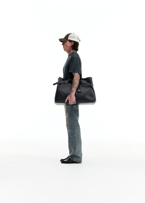 A man in a side profile wearing a baseball cap, dark t-shirt, and jeans, carrying a large black leather tote bag against a solid white background.