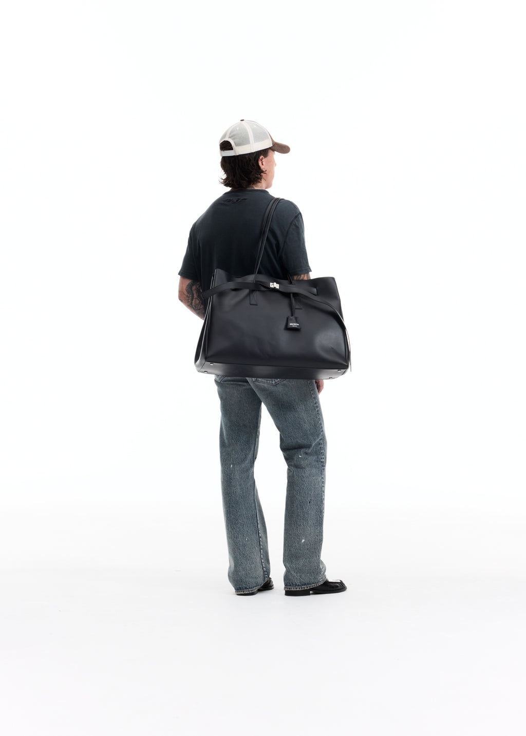 A man viewed from behind wearing a black t-shirt, blue jeans, and a white cap, carrying a large black leather duffel bag over his shoulder against a solid white background.