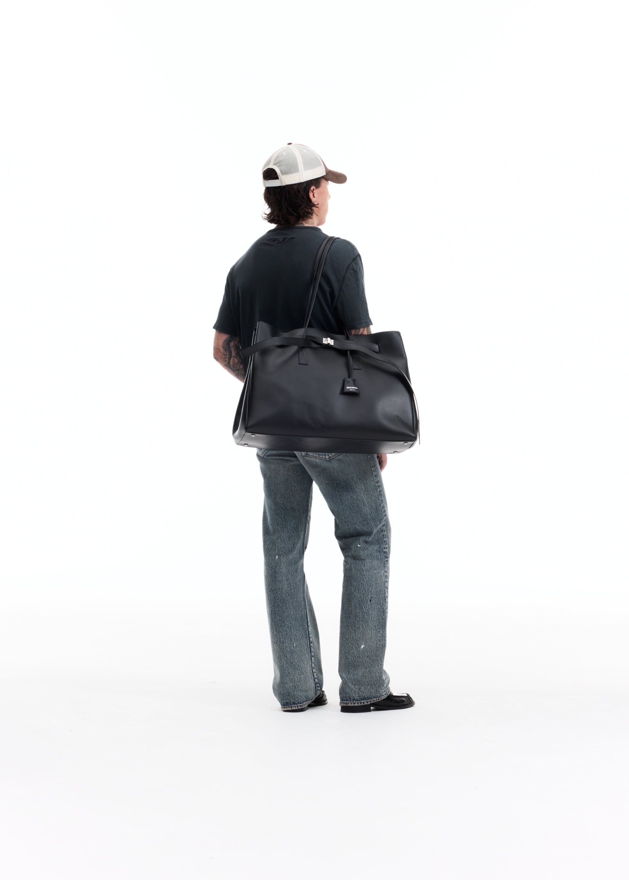 A man viewed from behind wearing a black t-shirt, blue jeans, and a white cap, carrying a large black leather duffel bag over his shoulder against a solid white background.
