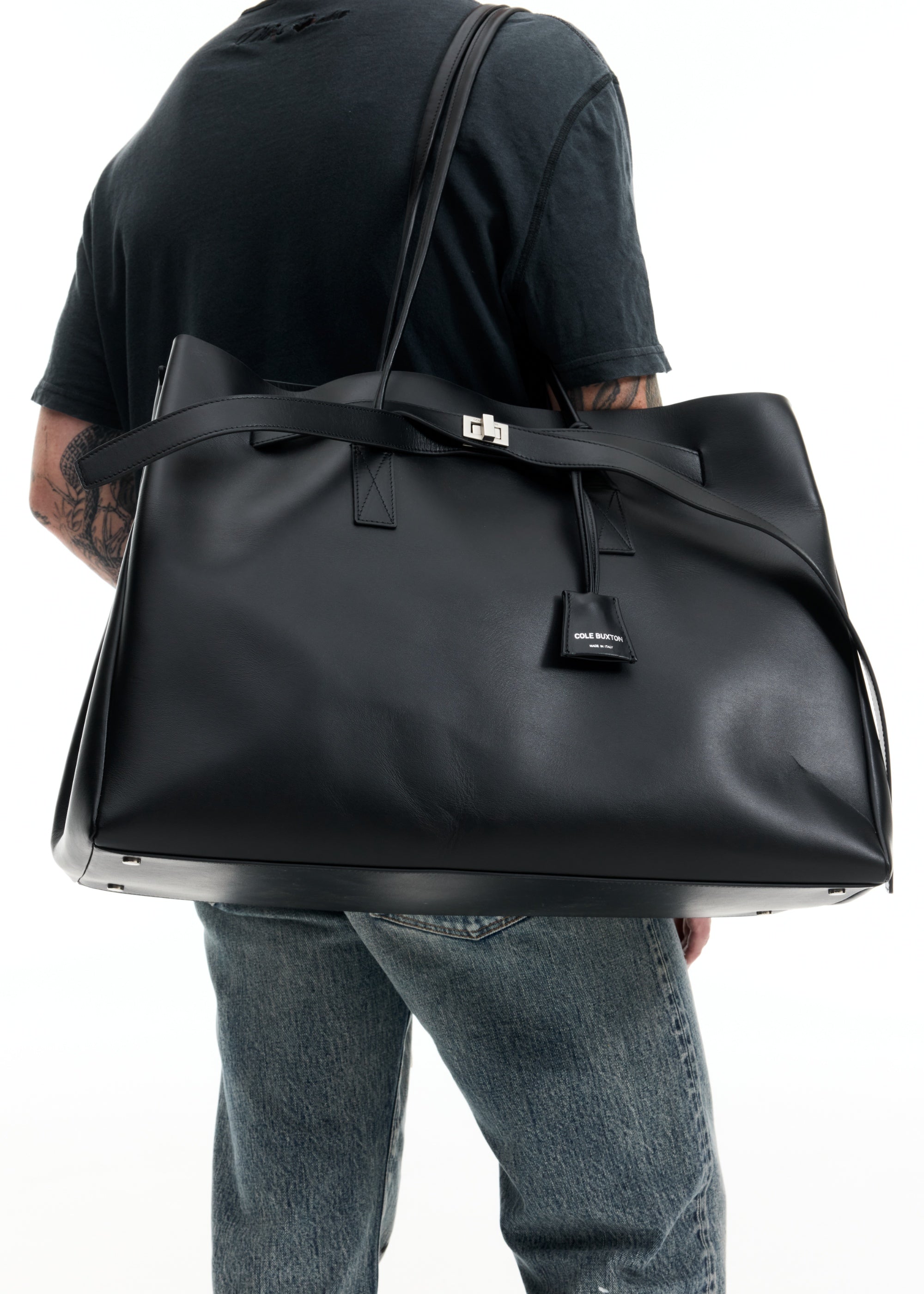 A person wearing a black t-shirt and blue jeans carries a large black leather tote bag over their shoulder against a white background.