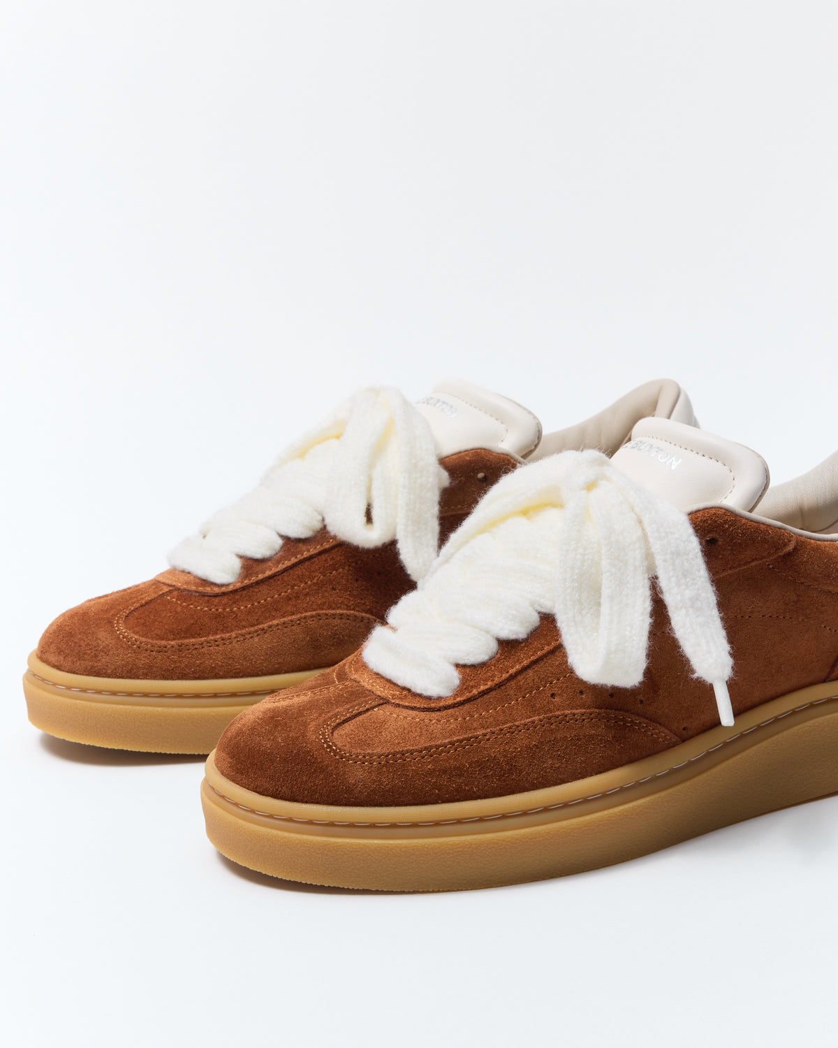 A pair of brown suede low-top sneakers with thick white faux-fur laces and white lining, featuring a gum rubber sole, photographed on a clean white studio background.