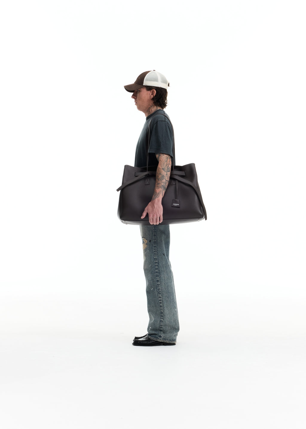 A man in a dark t-shirt, jeans, and a trucker hat stands in profile holding a large black leather duffel bag against a solid white studio background.