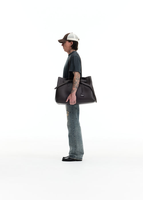 A man in a dark t-shirt, jeans, and a trucker hat stands in profile holding a large black leather duffel bag against a solid white studio background.
