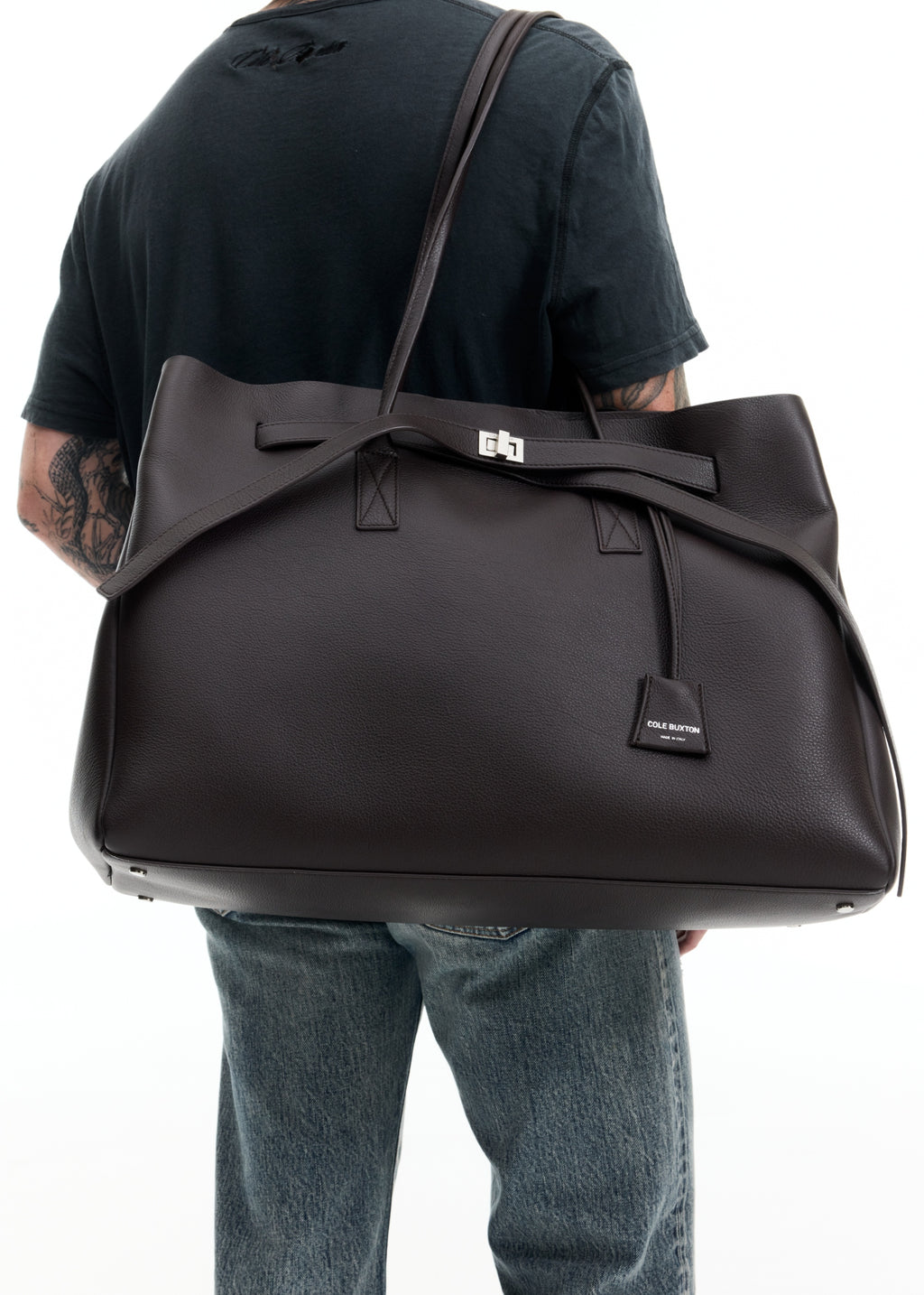 A person wearing a black t-shirt and blue jeans carries a large black leather duffel bag with a structured top handle and silver hardware against a white background.