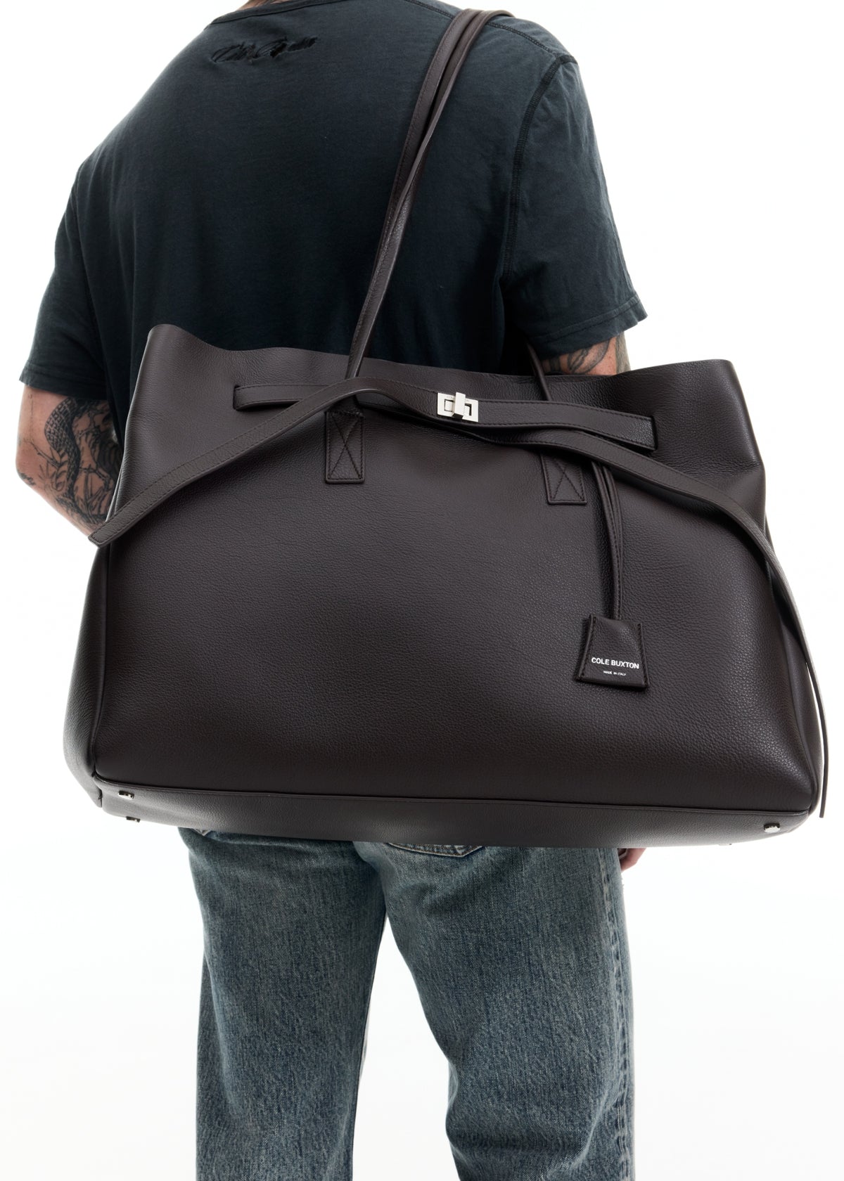 A person wearing a black t-shirt and blue jeans carries a large black leather duffel bag with a structured top handle and silver hardware against a white background.