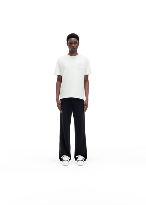 A man modeling a white short-sleeve t-shirt with a subtle circular logo on the left chest, paired with black wide-leg trousers and white sneakers, shot against a seamless white background.