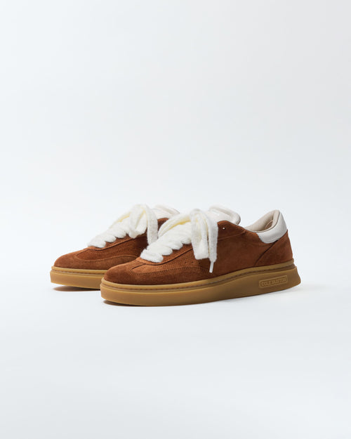 A pair of brown suede low-top sneakers with thick white faux-fur laces and white lining, featuring a gum rubber sole, photographed on a clean white studio background.