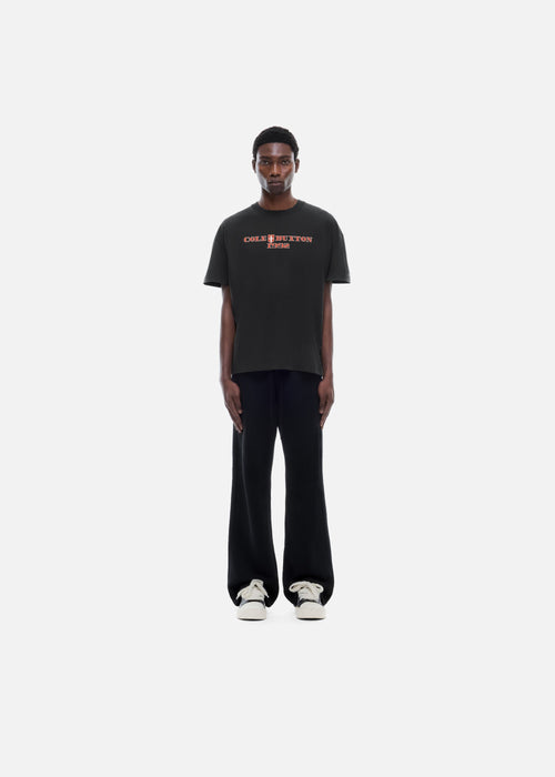 A model stands against a plain light gray background wearing a black short-sleeve T-shirt with orange-red text reading 'CHARLIE BROWN PARIS' across the chest, black wide-leg pants, and white sneakers.
